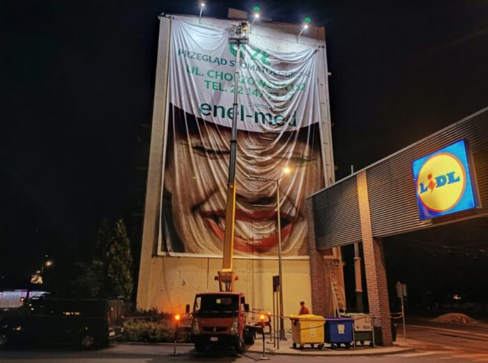 Alpiniści montujący reklamę z podnośnika koszowego, prace montażowe na wysokości, instalacja reklamy na elewacji budynku.